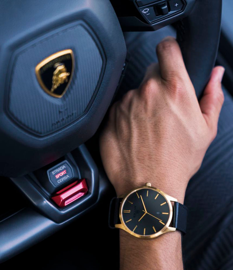 Reloj para hombre marca Bellum, color oro amarillo 14k en acabado cepillado con dial negro mate y correa de cuero negro. En muñeca de hombre agarrando volante de Lamborghini Huracán.