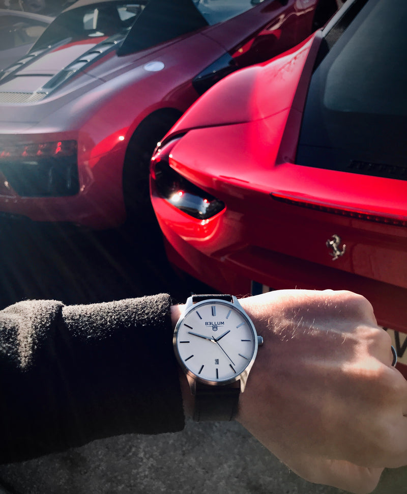 Reloj para hombre marca Bellum, color plata con dial blanco y correa de cuero negra. Vista frontal en muñeca de hombre con Ferrari 458 y Audi R8.